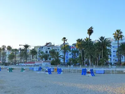 a beach with blue chairs and palm trees and buildings at Sitges Center & Beach with Sea View in Sitges