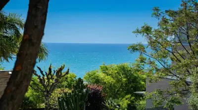 a view of the ocean from behind some trees at Villa Sitges Colibri at 10 min Walk Beaches - Center City Amaizing Garden Pool XXL Private Tenis Piste in Vilanova i la Geltrú